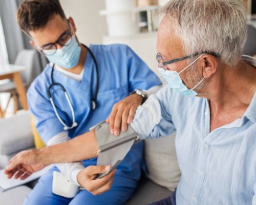 Male nurse measures blood pressure to senior man with mask while being in a home visit.